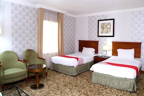 Habitación de hotel con dos camas, una mesita pequeña, dos sillones, una ventana con cortinas y un cuadro en la pared.