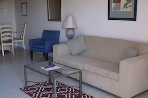 Un salon avec un canapé beige, un fauteuil bleu, une table basse en verre et un tapis à motifs.