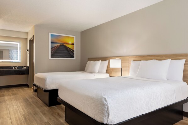 A hotel room with two beds, a wooden headboard, a painting of a sunset, a bathroom with a mirror and sink, and a wooden floor. - Days Inn by Wyndham Kuttawa/eddyville (Kuttawa)