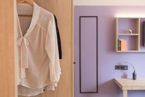 A room with a wooden wardrobe, a hanging garment, a purple wall, a framed mirror, a shelf with books, and a small table with a vase. - Yggotel Solsort (Essen)