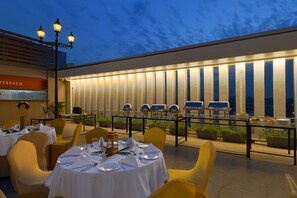 Un área de comedor al aire libre con mesas dispuestas para cuatro, un bar exterior con varias parrillas y vistas del horizonte de la ciudad al atardecer.
