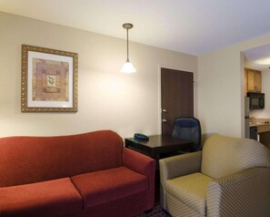 Habitación de hotel con un sofá rojo, un sillón estampado, un escritorio con silla, un cuadro enmarcado en la pared y una zona de cocina.