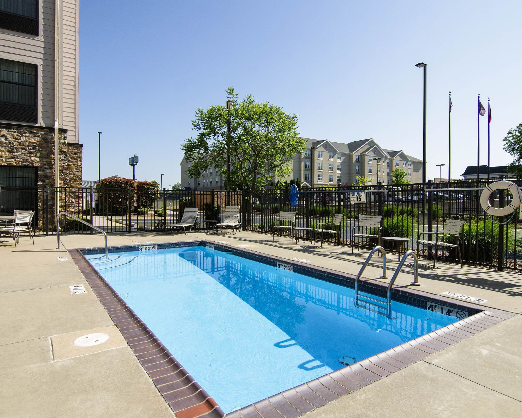 A rectangular swimming pool with a metal ladder, surrounded by a black fence, and a stone building to the left.