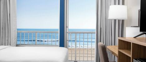 A hotel room with a bed, a desk, a chair, a TV, and a balcony with a view of the sea.