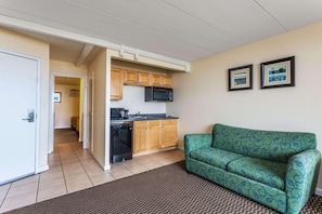 A compact living space with a kitchenette, a green patterned sofa, and a door leading to another room. - Travelodge by Wyndham Suites Virginia Beach Oceanfront (Virginia Beach)