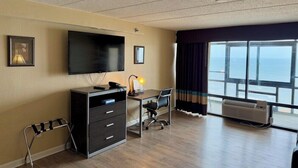 A hotel room with a large TV, a desk with a chair, a dresser, a window with curtains, and a view of the ocean. - Travelodge by Wyndham Suites Virginia Beach Oceanfront (Virginia Beach)