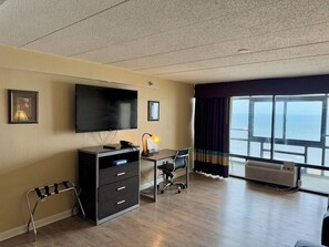 Habitación de hotel con un televisor grande, un escritorio con silla, cómoda, ventana con cortinas y vista al océano.