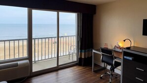 A hotel room with a large sliding glass door to the beach, a desk with a chair, and a small table with a lamp. - Travelodge by Wyndham Suites Virginia Beach Oceanfront (Virginia Beach)