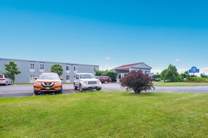 A parking lot with several cars, a building with a red roof, and a blue sign with a white symbol. - Motel6 Oriskany, NY (Oriskany)