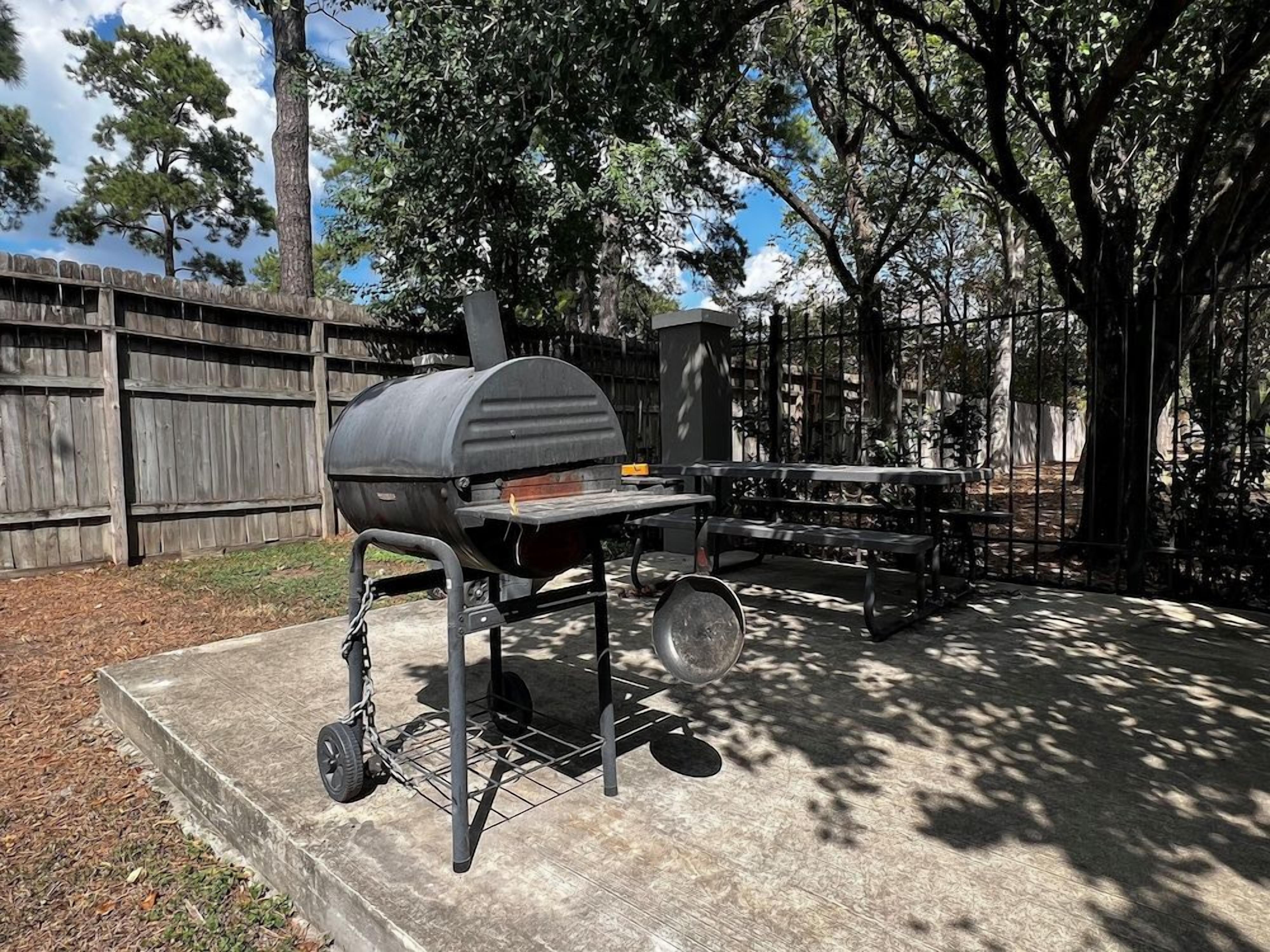 A barbecue grill with a chimney, a table, and a hanging pan, set in a backyard with a wooden fence and trees.