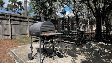A barbecue grill with a chimney, a table, and a hanging pan, set in a backyard with a wooden fence and trees.