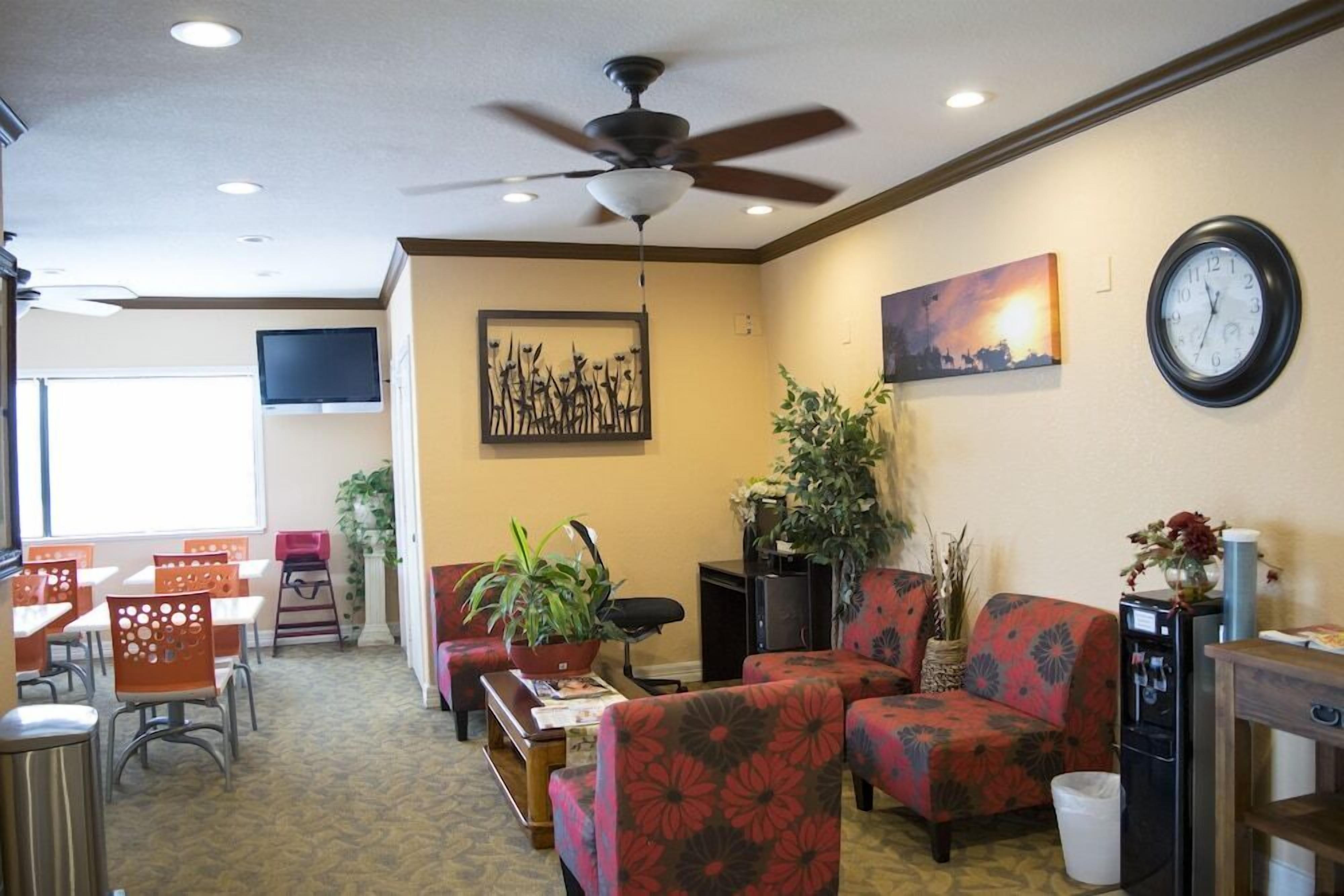 A hotel lobby with a ceiling fan, a clock, a painting, and a seating area with chairs and tables.