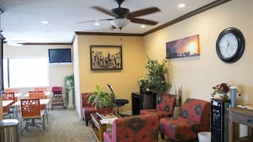 A hotel lobby with a ceiling fan, a clock, a painting, and a seating area with chairs and tables.