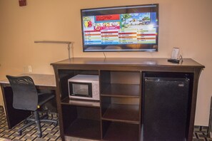 A hotel room with a TV mounted on the wall, a microwave in a cabinet, a refrigerator, and a desk with a chair.