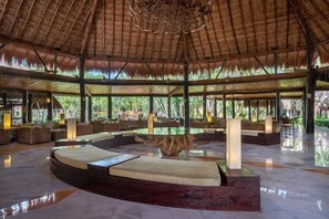 Un vaste espace intérieur doté d’un haut plafond, une table centrale en verre entourée de sièges, et une vue sur une végétation luxuriante à l’extérieur.