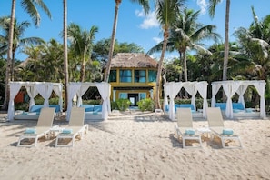 Un resort en la playa con un edificio de techo de paja, zonas de estar con dosel blanco y palmeras.
