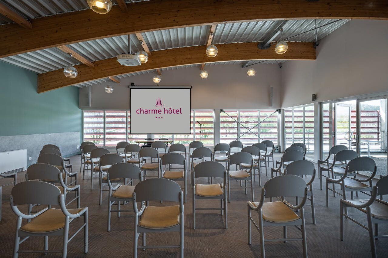 Un salón de conferencias con filas de sillas, una pantalla de proyector y un ventanal con vistas al exterior.