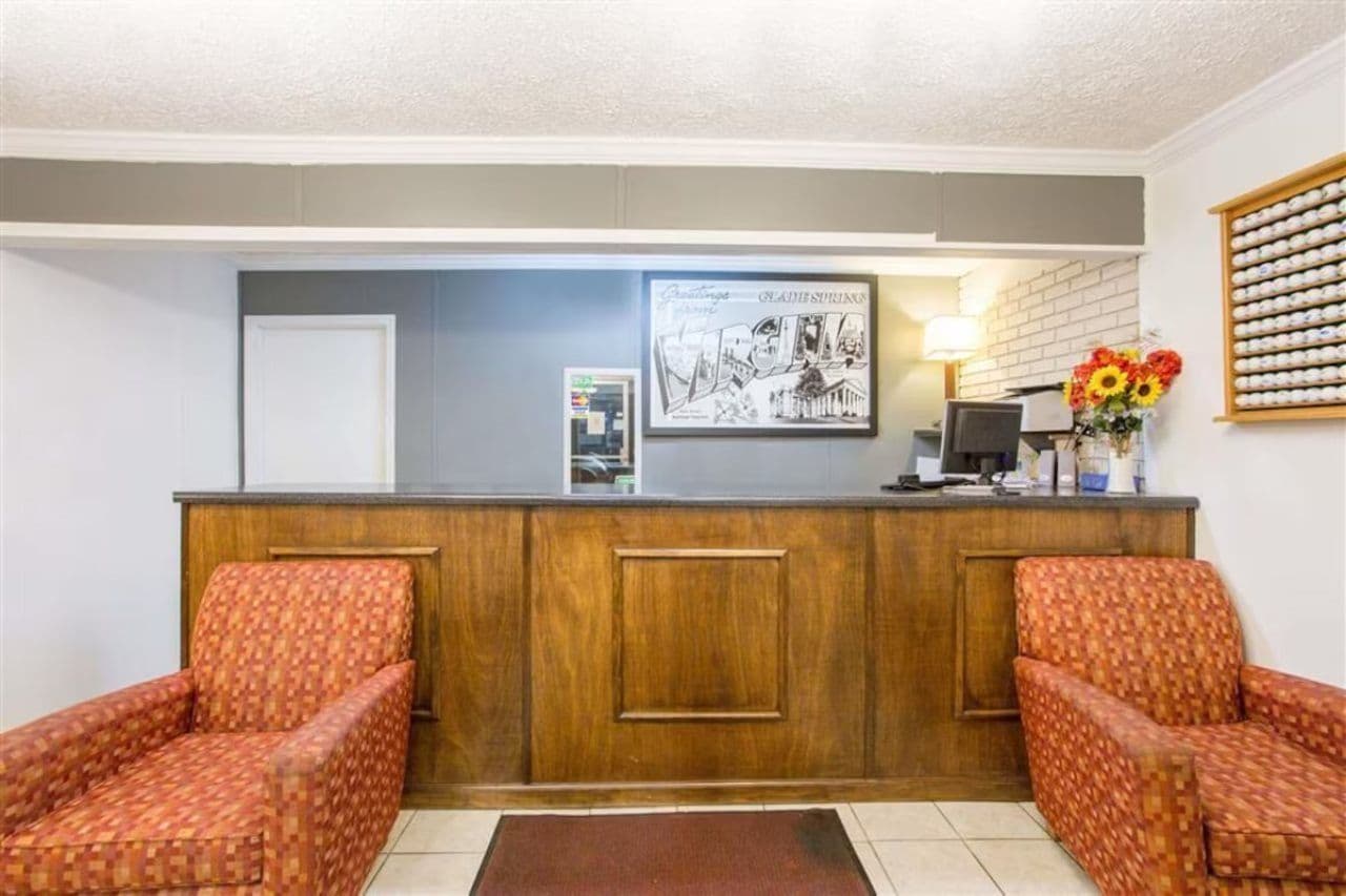 A reception area with a wooden desk, two patterned armchairs, a framed picture, a vase with flowers, and a wall-mounted clock.