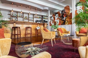 A bar area with stools, a counter with bottles, and a mural. There are chairs and a table with a decorative item. - The Originals City, Hotel Bristol, Le Puy-en-velay (Le Puy-en-Velay)