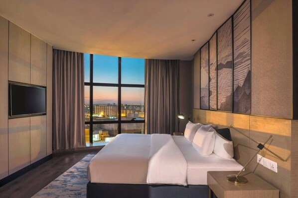A hotel room with a large bed, a flat-screen TV, a city view through the window, and a modern design. - Aber Unaiza (Unaizah)