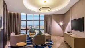A modern hotel room with a large window, a round ceiling light, a flat-screen TV, and a seating area with a geometric patterned rug. - Aber Unaiza (Unaizah)