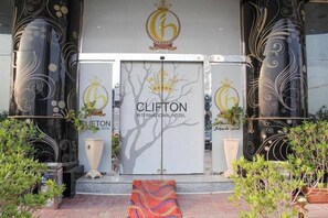 Entrada do Hotel Clifton International, com uma placa grande, colunas decorativas e vasos de plantas.