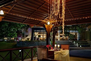 Un área de comedor al aire libre con techo de paja, luces colgantes y una mesa preparada para dos.