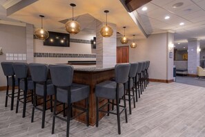 Uma área de bar com balcão alongado e vários banquetas, uma televisão na parede e iluminação pendente.