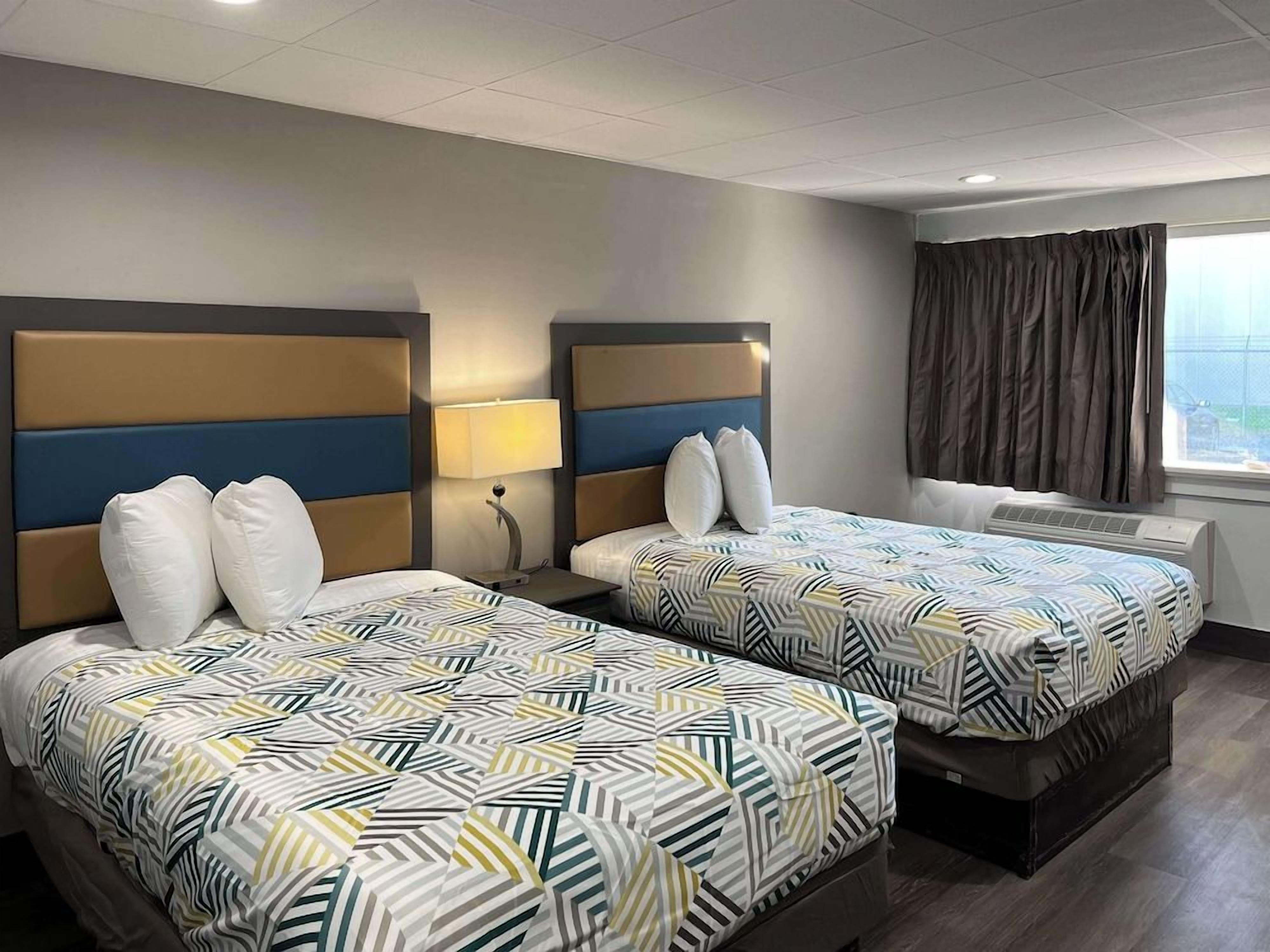 A hotel room with two beds, a window with curtains, and a wall-mounted air conditioning unit.