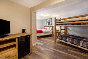 Habitación de hotel compacta con una litera de madera, un escritorio pequeño con televisor y cafetera, y una cama con manta escocesa.