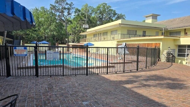 A swimming pool surrounded by a brick patio, a fenced area, and a yellow building with a balcony.