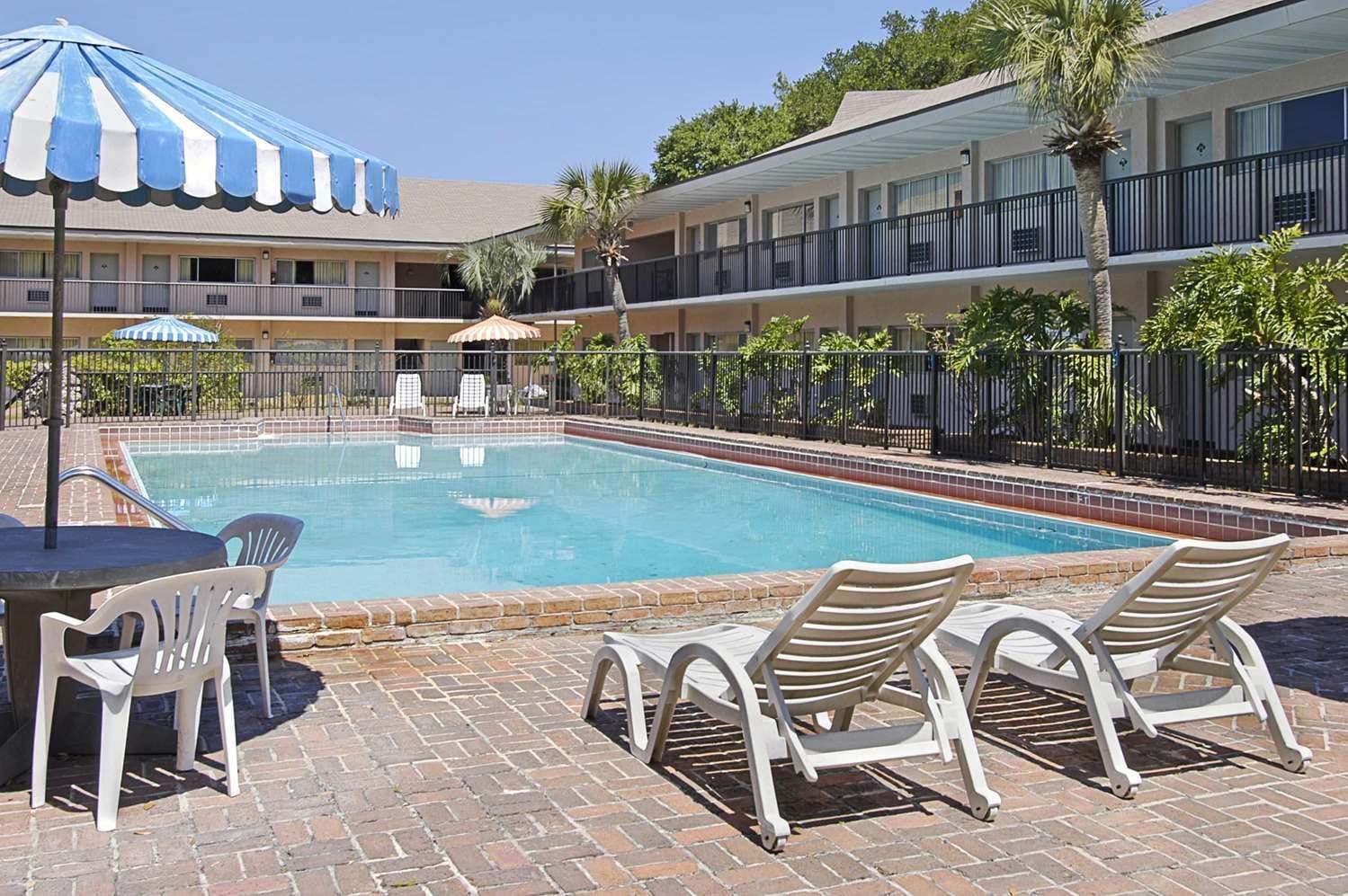 Une piscine entourée de chaises longues et de parasols, elle-même bordée d’une terrasse en briques et d’un immeuble à plusieurs étages doté de balcons.