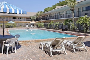 Piscina com espreguiçadeiras e guarda-sóis, rodeada por um terraço de tijolos e um edifício com varandas.