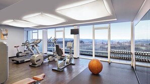 A gym with exercise equipment, a large orange exercise ball, and a view of the city through large windows. - Lindner Hotel Nurburgring Congress (Nuerburg)