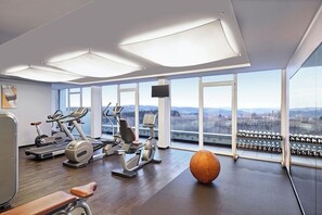 Ein Fitnessstudio mit Trainingsgeräten, einem großen orangefarbenen Trainingsball und Blick auf die Stadt durch große Fenster.