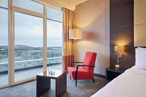 Una habitación de hotel con un ventanal grande, una silla roja, una mesita de noche, una lámpara y una cama.