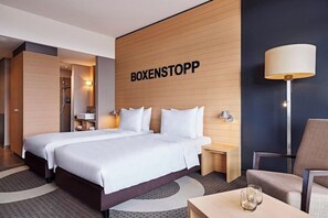 A hotel room with two beds, a wooden accent wall, a lamp, a chair, and a small table. - Lindner Hotel Nurburgring Congress (Nuerburg)