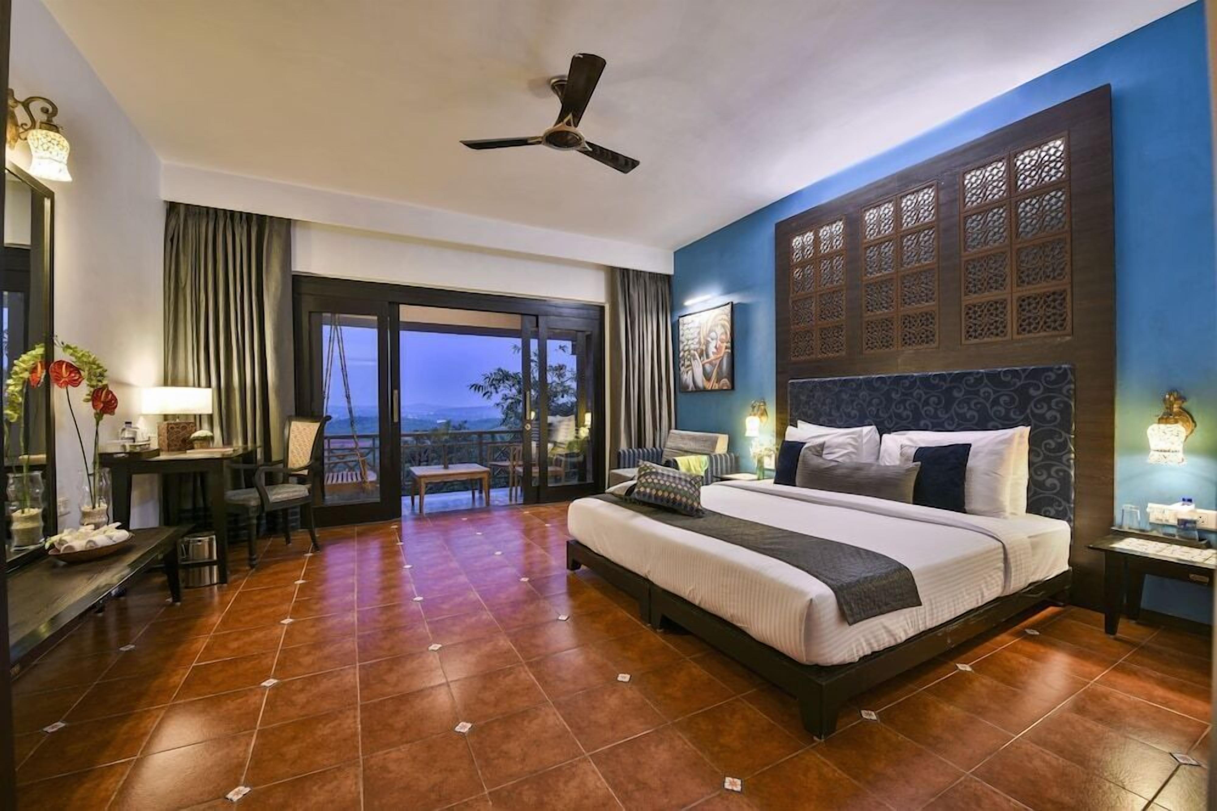 A hotel room with a large bed, a ceiling fan, a balcony with a view, and a dining area with a table and chairs.