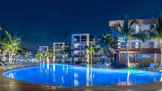 A swimming pool with lounge chairs and palm trees, surrounded by modern buildings. - Blue Beach Punta Cana Luxury Resorts (Punta Cana)