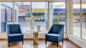 A room with two blue armchairs, a small table with a vase of flowers, and large windows offering a view of the street.