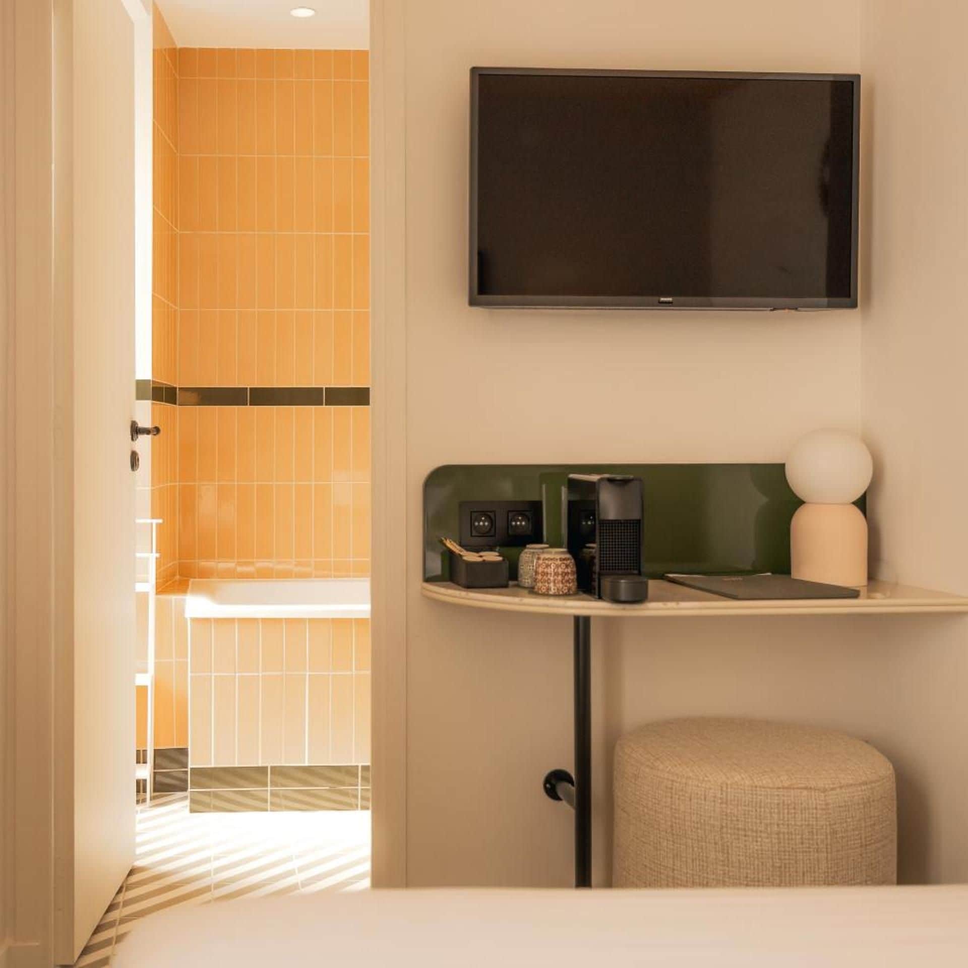 A hotel room with a flat-screen TV mounted on the wall, a small table with a lamp, and a seating area with a cushioned stool.