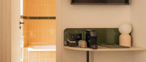A hotel room with a flat-screen TV mounted on the wall, a small table with a lamp, and a seating area with a cushioned stool.