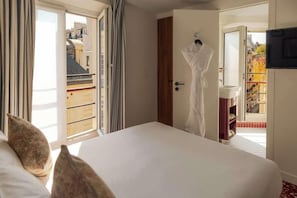 Ein Hotelzimmer mit Bett, einem an der Tür hängenden Bademantel, Blick auf Gebäude und einem Balkon mit Geländer.