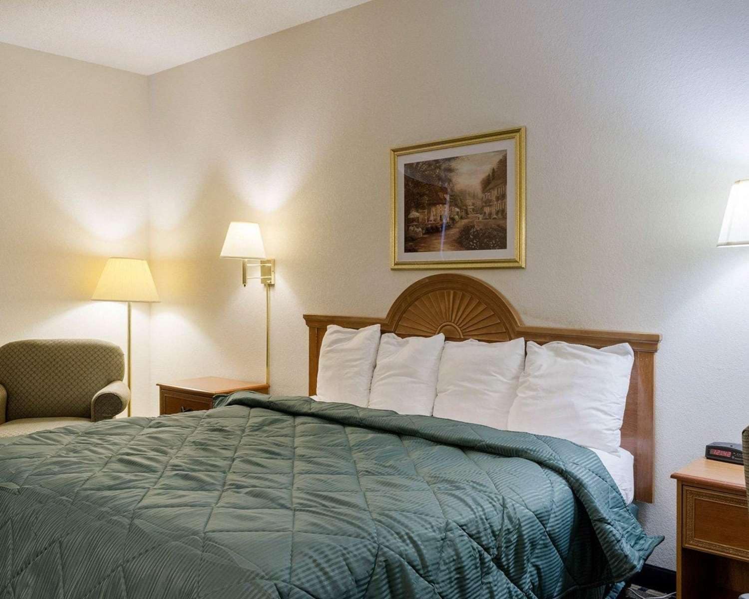 A hotel room with a bed, a chair, a nightstand, a lamp, and a painting on the wall.