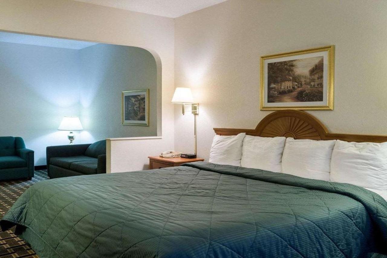 A hotel room with a large bed, a nightstand, a lamp, a chair, and a painting on the wall.