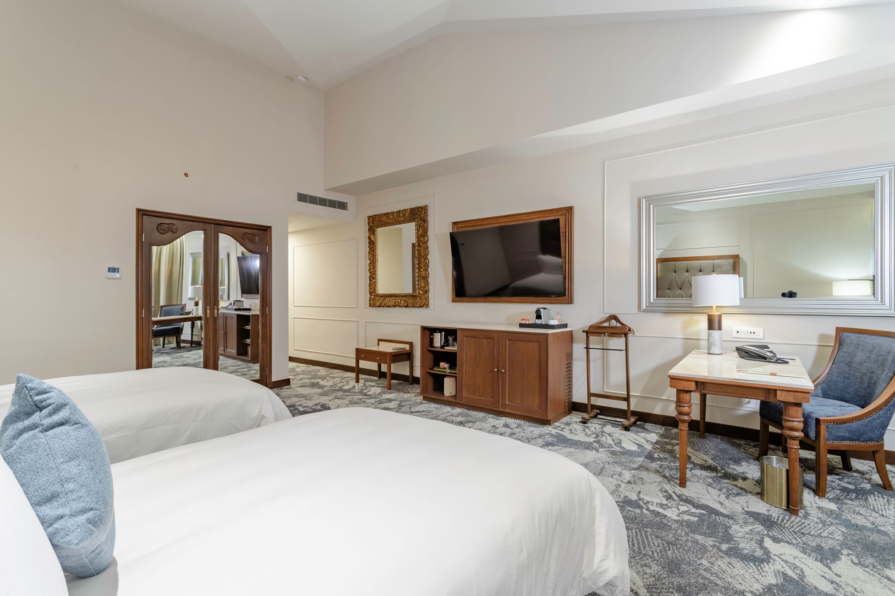 Habitación de hotel con dos camas, un escritorio, una silla, un televisor y un espejo grande.