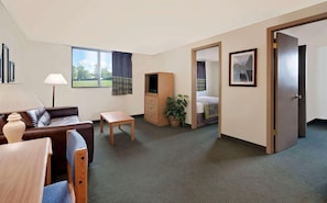 Ett hotellrum med en soffa, ett litet bord, ett TV-bord, en säng, en dörr och ett fönster med utsikt över ett grönskande landskap.