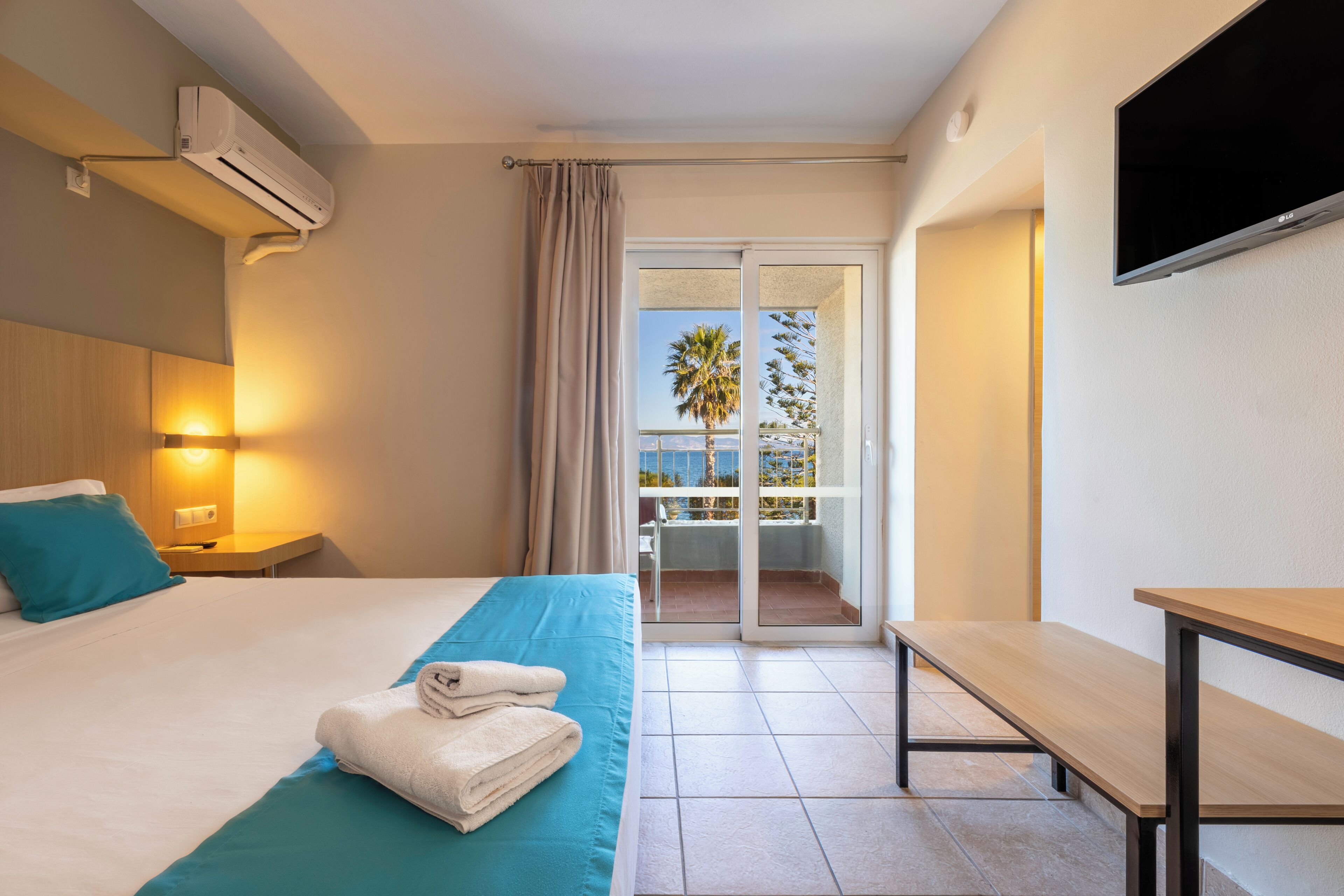 Habitación de hotel con cama, una banca, televisión y balcón con vistas.