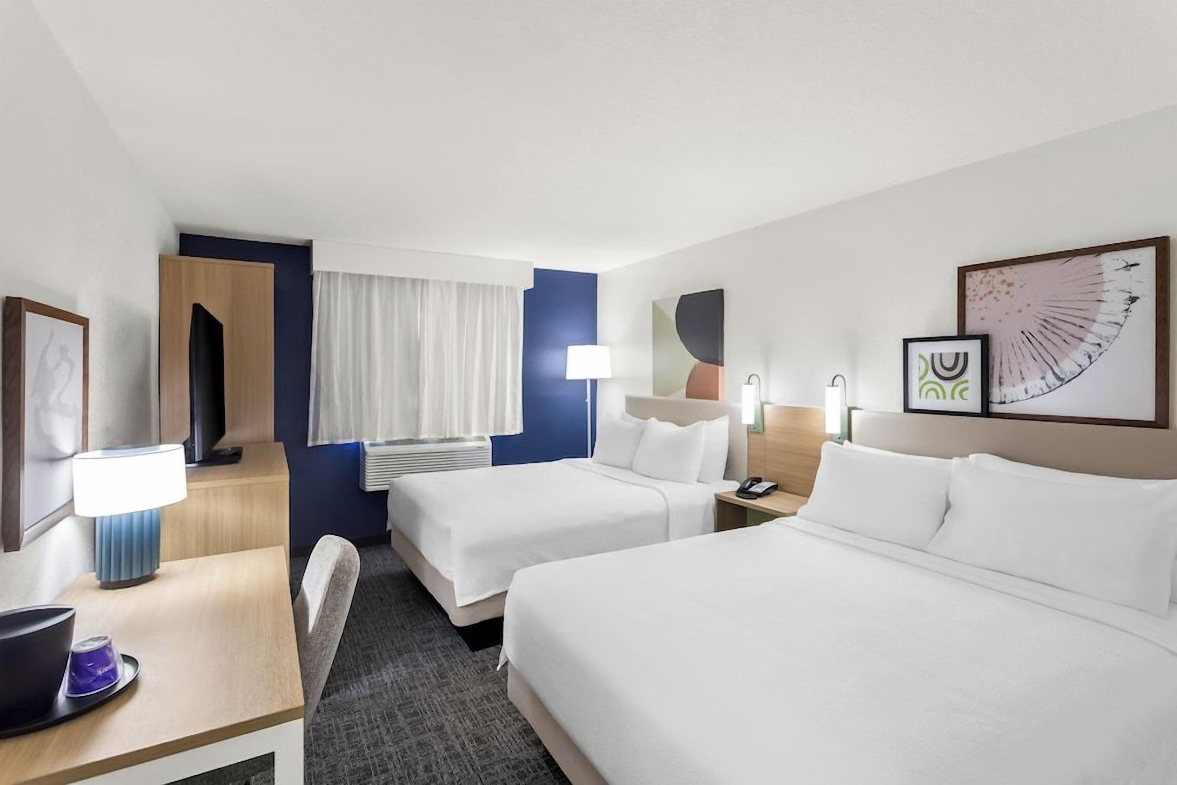 A hotel room with two beds, a desk with a lamp, a chair, and artwork on the walls.
