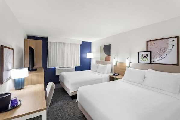 Habitación de hotel con dos camas, un escritorio con lámpara, una silla y obras de arte en las paredes.